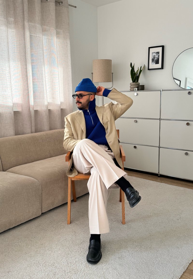 Blazer beige sur un col roulé bleu, pantalon blanc plissé, chaussures noires. Assis sur une chaise en bois avec un canapé gris clair et une plante en arrière-plan.