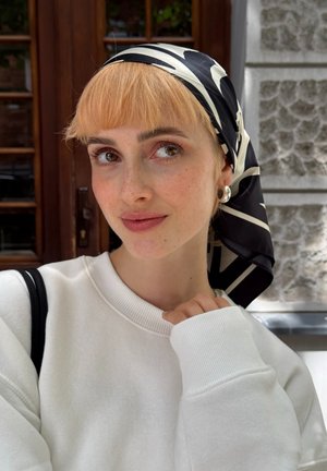 Bufanda de seda con patrón en blanco y negro, sudadera blanca suave con puños acanalados. La persona tiene el cabello corto de color rubio claro y aretes de aro plateados.