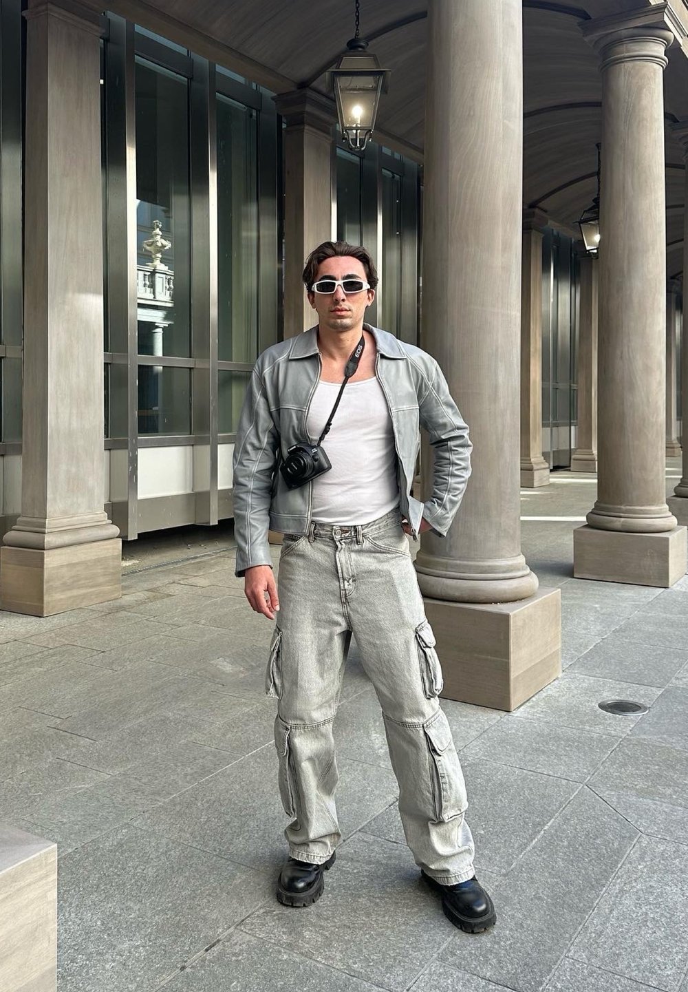 Ανοιχτό γκρίζο δερμάτινο μπουφάν πάνω από ένα λευκό τοπ, συνδυασμένο με φαρδιά γκρίζα cargo παντελόνια και μαύρες μπότες. Η κάμερα κρέμεται από τον λαιμό, φοράει γυαλιά ηλίου.