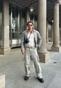 Ανοιχτό γκρίζο δερμάτινο μπουφάν πάνω από ένα λευκό τοπ, συνδυασμένο με φαρδιά γκρίζα cargo παντελόνια και μαύρες μπότες. Η κάμερα κρέμεται από τον λαιμό, φοράει γυαλιά ηλίου.