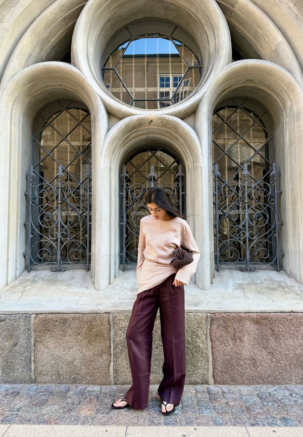 Femme en haut rose clair et pantalon foncé, pieds nus, debout sur une rue pavée devant des fenêtres voûtées ornées avec des grilles en fer noir.