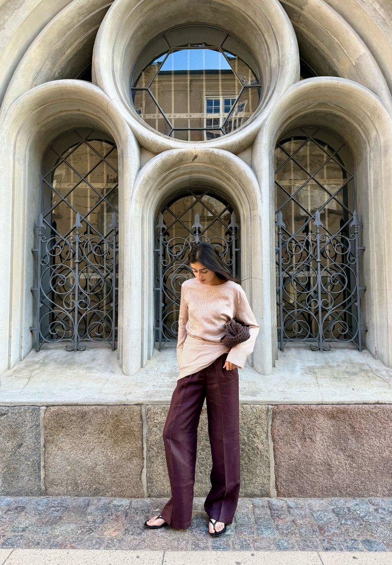 Femme en haut rose clair et pantalon foncé, pieds nus, debout sur une rue pavée devant des fenêtres voûtées ornées avec des grilles en fer noir.