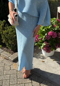 Vestido azul-claro plissado com pontos brancos dispersos, bainha com borda recortada, combinado com sapatos de salto cor-de-rosa e uma bagagem branca texturizada. Plantas verdes ao fundo.