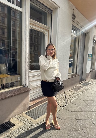 Suéter beige claro con mangas texturizadas, falda corta negra y bolso de mano negro trenzado. De pie en la acera con chanclas, cerca de una tienda.