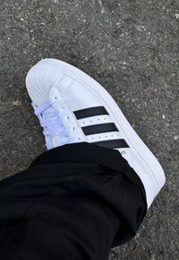 Λευκό αθλητικό παπούτσι με μαύρες ρίγες φορεμένο σε πόδι πάνω σε τραχύ άσφαλτο, σε συνδυασμό με μαύρο παντελόνι.