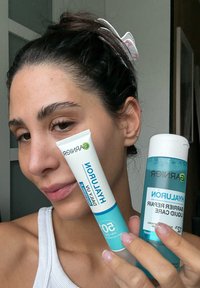 Kvinne med mørkt hår i en hvit topp som holder Garnier Hyaluron ansiktskrem og væskepleieprodukter nær ansiktet.