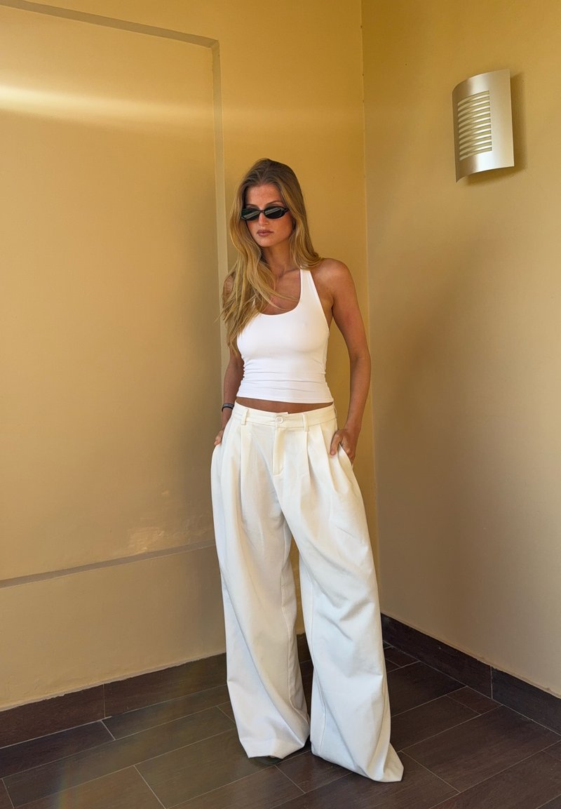 Γυναίκα με μακριά ξανθά μαλλιά, φοράει μαύρα γυαλιά ηλίου, λευκό αμάνικο τοπ και λευκό παντελόνι με φαρδιά πόδια, στέκεται μπροστά σε μπεζ τοίχο.