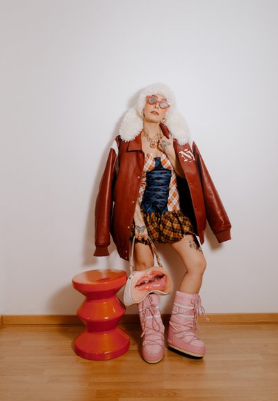 Chaqueta bomber de cuero rojo con cuello de piel, top corsé azul, falda de cuadros, botas rosas hasta la rodilla, bolso de cubo rosa con diseño de labios y taburete rosa.