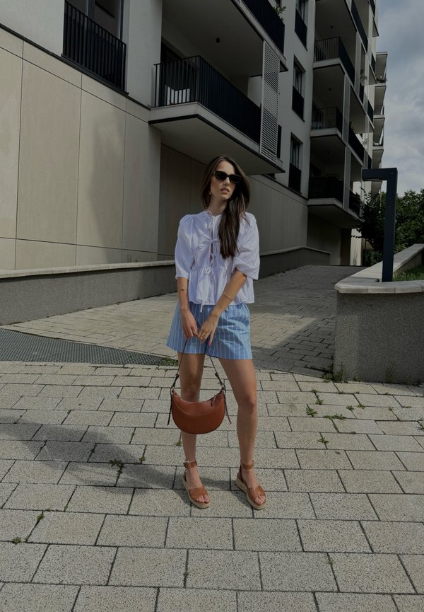 Blusa bianca a maniche corte con maniche arricciate, pantaloni corti a righe blu, borsa marrone e sandali marroni con cinturini, in piedi su un terreno pavimentato.