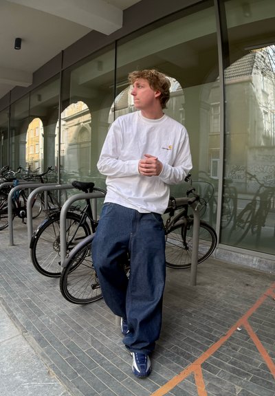 Camisa blanca de manga larga con logo, pantalones vaqueros azules holgados y zapatillas deportivas oscuras. Apoyado contra un soporte de bicicletas con bicicletas al fondo.