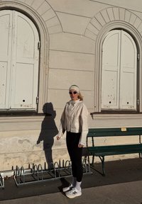 Veste légère de couleur crème avec un col montant et des manches longues ; leggings noirs ajustés ; baskets blanches. En arrière-plan, un mur beige et un banc.