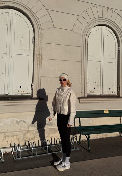 Chaqueta ligera color crema con cuello alto y mangas largas; leggings negros ajustados; zapatillas blancas. El fondo presenta una pared beige y un banco.