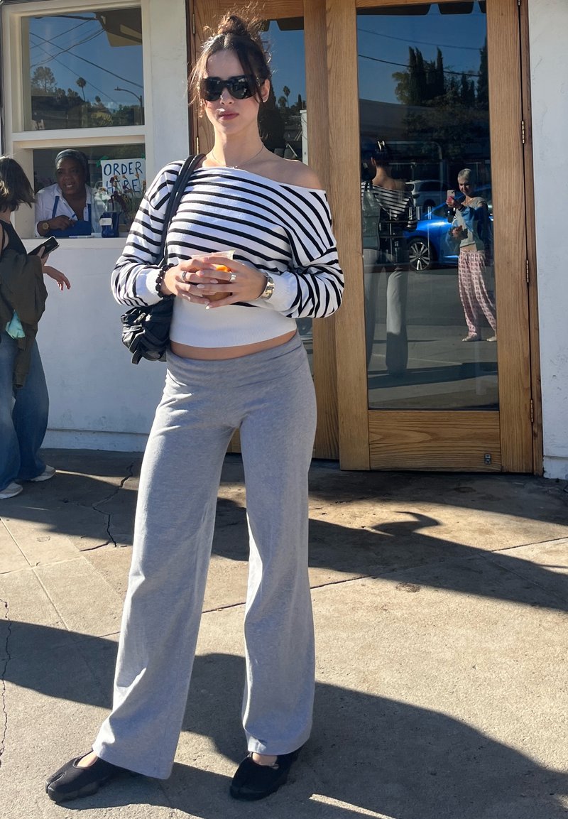 Striped off-shoulder jumper, grey joggers, black shoes. Woman holding a drink, standing outside a building with a wooden door.