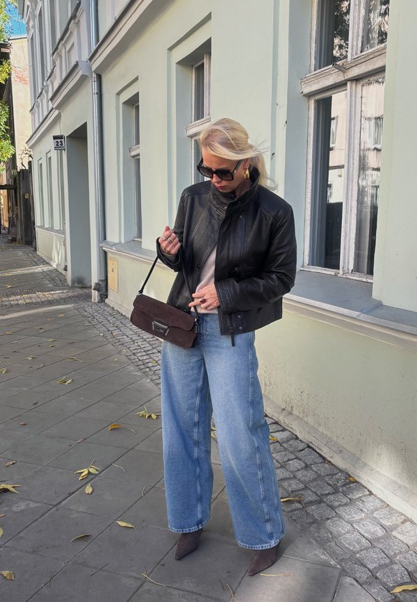 Svart läderjacka, grå topp, vida blå jeans och grå ankleboots, som håller en brun mockaväsk, stående på en trottoar.
