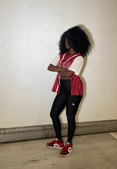 Jersey a rayas rojas y blancas con detalle de botones, leggings negros y zapatillas rojas y negras. Cabello rizado y negro. Fondo neutro.