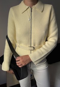 Persoană îmbrăcată cu o jachetă tricotată de un galben pal, cu baretă și fermoar, pantaloni albi, ținând o geantă neagră și purtând un colier cu pandantiv din argint.