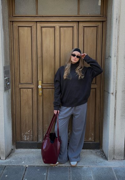 Suéter negro de gran tamaño con cuello alto, pantalones de pierna ancha a rayas grises y un bolso tote burdeos, frente a un fondo de puerta de madera.