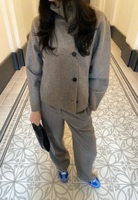 Pelēka vilnas maisījuma jaka ar augstu apkakli un pogu aizdari, kombinēta ar pelēkiem biksēm un zilo un baltu sporta apavu pāri. Melna sajūga soma rokā.