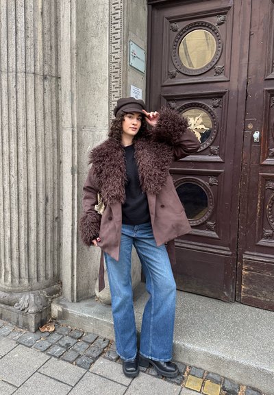 Abrigo marrón con borde de piel sintética, diseño de doble botonadura, combinado con suéter negro y jeans azul de pierna ancha, posando frente a una puerta de madera.