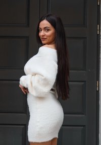 Femme aux longs cheveux foncés portant une robe blanche duveteuse à épaules dénudées, debout de profil devant une porte à panneaux sombre, souriant doucement.