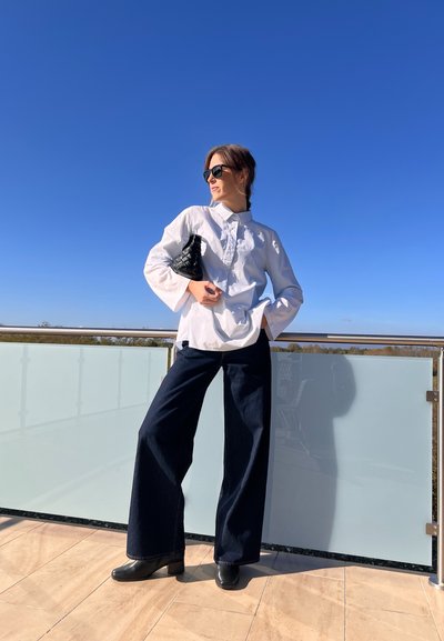 Camisa blanca con detalle de botones, pantalones de mezclilla oscuros y anchos, botas de tacón negras y un bolso negro texturizado en un ambiente exterior brillante.