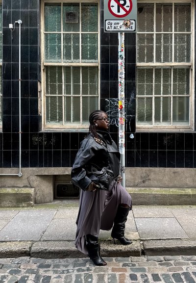Mujer con el cabello trenzado, vestida con una chaqueta de cuero negra, falda larga, botas y gafas de sol, caminando junto a una señal de no estacionar en una calle adoquinada.