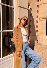 Tan suede lange jas over een witte top, gecombineerd met lichtblauwe wijde jeans. Zwarte handtas met gouden hardware. Zonnebril maakt de look compleet.