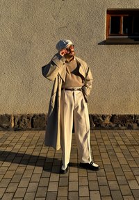 Sobretudo bege sobre um suéter claro, calças creme e sapatos preto e branco. Os destaques incluem um chapéu estilo balde e óculos.