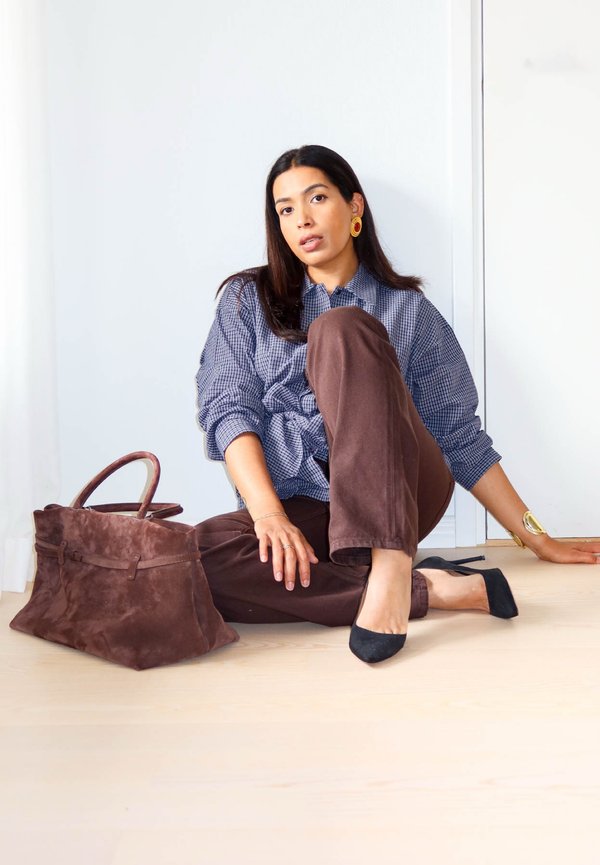 Mujer sentada en el suelo con pantalones marrones, camisa de cuadros, tacones negros, joyas de oro y un gran bolso de ante marrón a su lado.