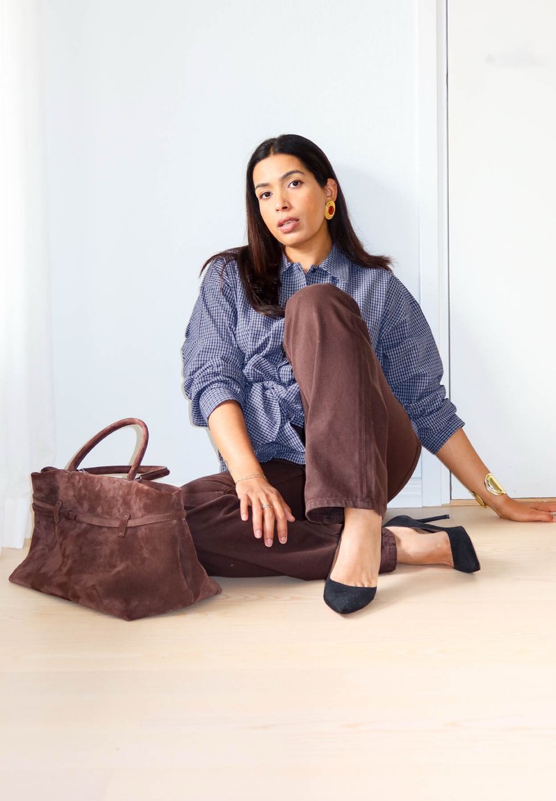 Mujer sentada en el suelo con pantalones marrones, camisa de cuadros, tacones negros, joyas de oro y un gran bolso de ante marrón a su lado.