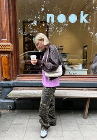 Jachetă bomber maro, tricou violet, pantaloni cu imprimeu leopard și adidași gri. Ținând o cană de cafea, o geantă albă texturată pe umăr, stând în aer liber.