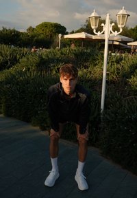 Črna atletska jakna z zadrgo, črne kratke hlače in bele športne čevlje. Stoji na kamniti poti ob zelenih grmovjih in zunanjem pohištvu.