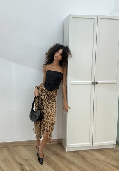 Top corsé negro sin tirantes, falda midi de estampado animal con drapeado, bolso negro elegante, tacones negros de punta, suelo de madera clara, fondo neutro.