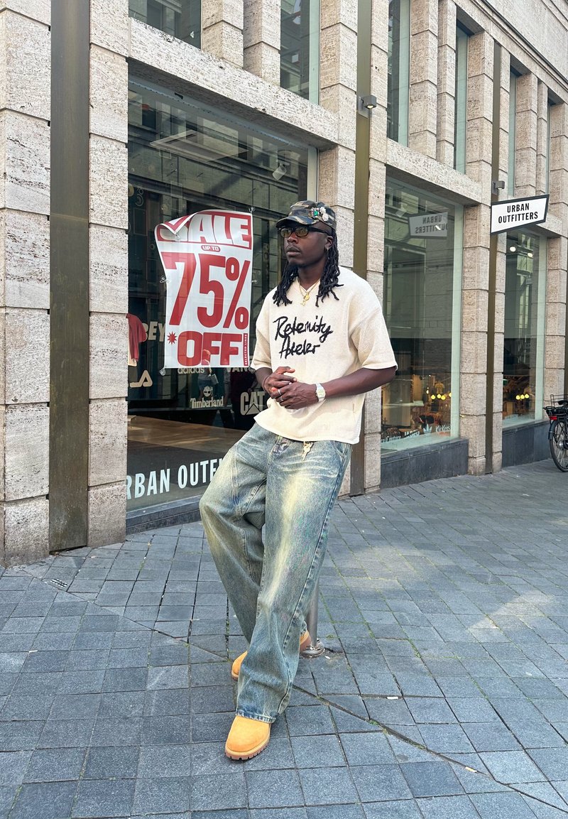 Beige oversize t-shirt med svarta bokstäver, ljusblå baggy jeans med en aning lyster, och orange mockasandlekskor utanför en urban butik.