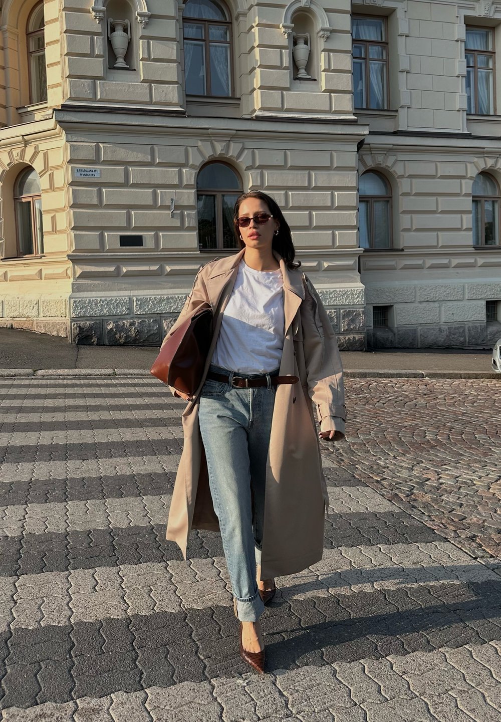 Μπεζ παλτό πάνω από λευκό πουκάμισο, μπλε τζιν και καφέ γόβες. Καφέ τσάντα, γυαλιά ηλίου, βαδίζει σε πλακόστρωτο.
