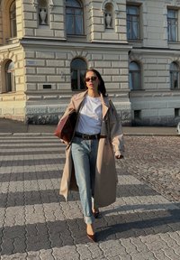 Μπεζ παλτό πάνω από λευκό πουκάμισο, μπλε τζιν και καφέ γόβες. Καφέ τσάντα, γυαλιά ηλίου, βαδίζει σε πλακόστρωτο.