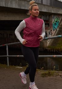 Burundu keskmise õmblusega vest valge pika varrukaga särgi peal, mustad retuusid ja roosad spordijalatsid. Jooksja kõnnib kanalile lähedasel asfalteeritud rajal.