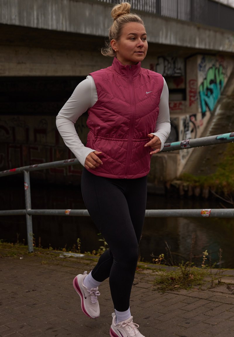 Bordinės spalvos siūtas liemenė ant balto ilgomis rankovėmis marškinėlių, juodos tamprės ir rožiniai sportiniai bateliai. Bėgikas asfaltuotu taku šalia kanalo.