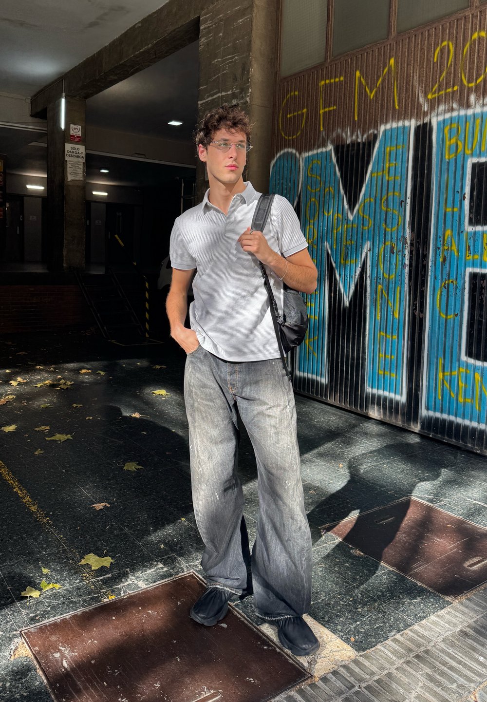 Polo gris à rayures, jeans gris amples, chaussures noires à enfiler, portant un sac bandoulière noir ; la lumière naturelle crée des ombres contrastées.