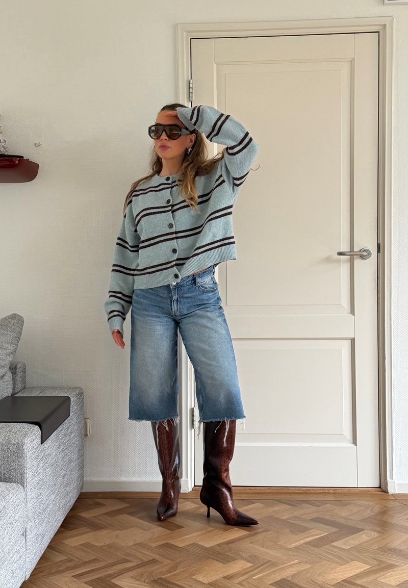 Personne portant un cardigan rayé bleu surdimensionné, un jean large court, des bottes à talons marron et de grandes lunettes de soleil noires, debout à l'intérieur.