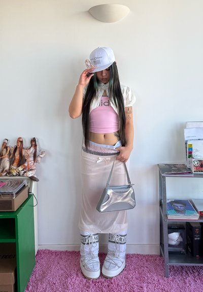 Joven con cabello largo, lleva una gorra blanca, top corto rosa, falda transparente, botas blancas y sostiene un bolso plateado en una habitación con estanterías y muñecas.