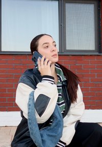 Personne aux cheveux longs tenant un smartphone noir, portant une veste bomber en cuir noir avec des manches blanches, une chemise rayée et un sac en denim.