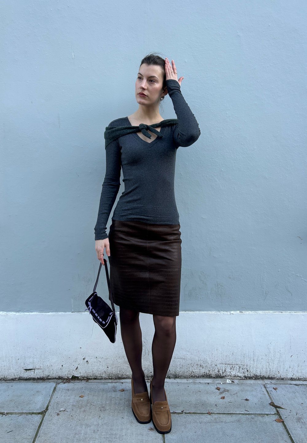 Haut gris à manches longues côtelé avec un col en V et un détail noué, associé à une jupe crayon en cuir marron. Accessoires comprenant un sac à main noir et des mocassins.