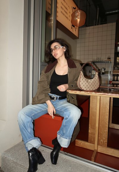Parka verde y marrón con cuello, camiseta sin mangas negra, jeans de mezclilla claros arremangados y botas de tacón negras. Bolsa de mano marrón con diseño sobre la mesa.
