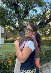 En kvinne iført store, gjennomsiktige grønne briller, en hvit skjorte, en mørk vest og et flerfarget skjerf, poserer i et gresskledd område.