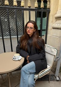 Pelēks vilnas mētelis pār melnu augšu, gaiši zilas džinsa bikses, caurspīdīgas brilles, sēžot pie apaļa bēša galda ar nelielu baltu kafijas krūzīti.