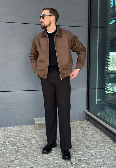 Chaqueta bomber de ante marrón con cuello, cierre de cremallera; suéter negro debajo, pantalones negros y calzado negro. Lleva gafas de sol cuadradas.