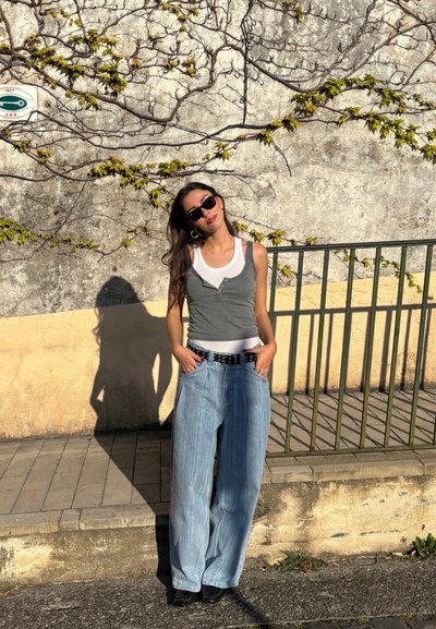 Top de tirantes gris ajustado sobre una camiseta blanca, combinado con jeans de pierna ancha a rayas azul claro y un cinturón negro. El fondo presenta una pared texturizada y enredaderas.