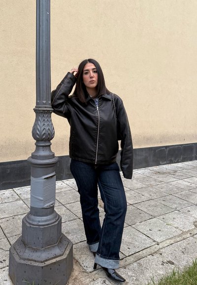 Chaqueta de cuero negra con cremallera, vaqueros de mezclilla azules con puños doblados y botas de tobillo puntiagudas negras, de pie junto a un farola.