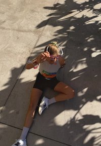 Tenue de sport avec un débardeur gris clair, un short noir et des baskets blanches. Porte des lunettes de soleil à verres roses, assis sur une surface en béton.